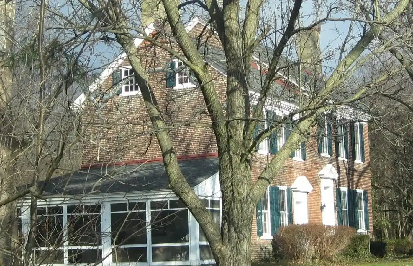 A historic two-story brick house in Gloucester County, featuring a screened-in porch and partially obscured by a large, leafless tree.