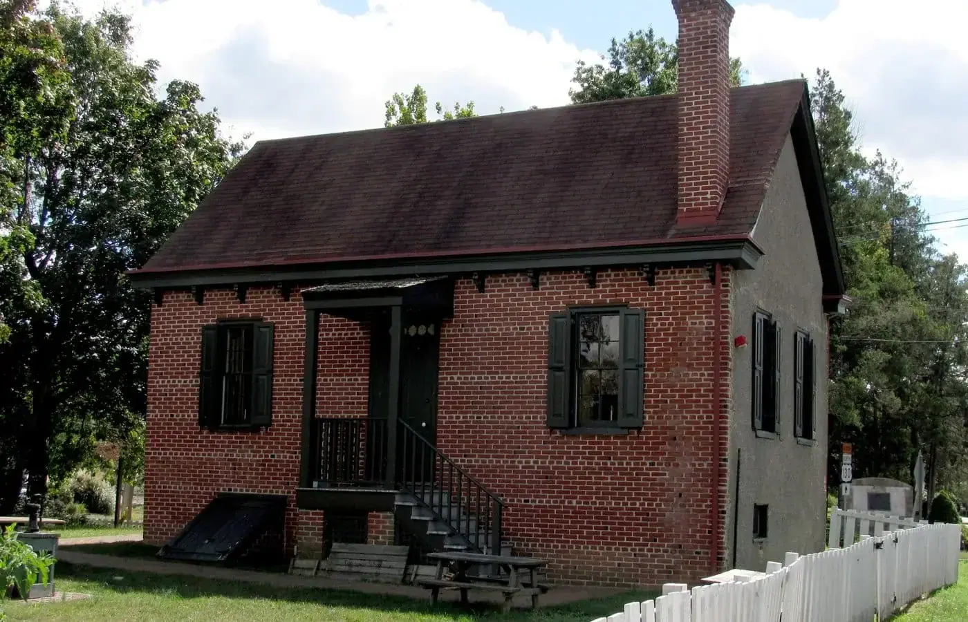 A historic red brick building in Camden County, featuring a dark shingled roof, a small front porch, and black shutters behind a white picket fence, expertly maintained by South Jersey painters.