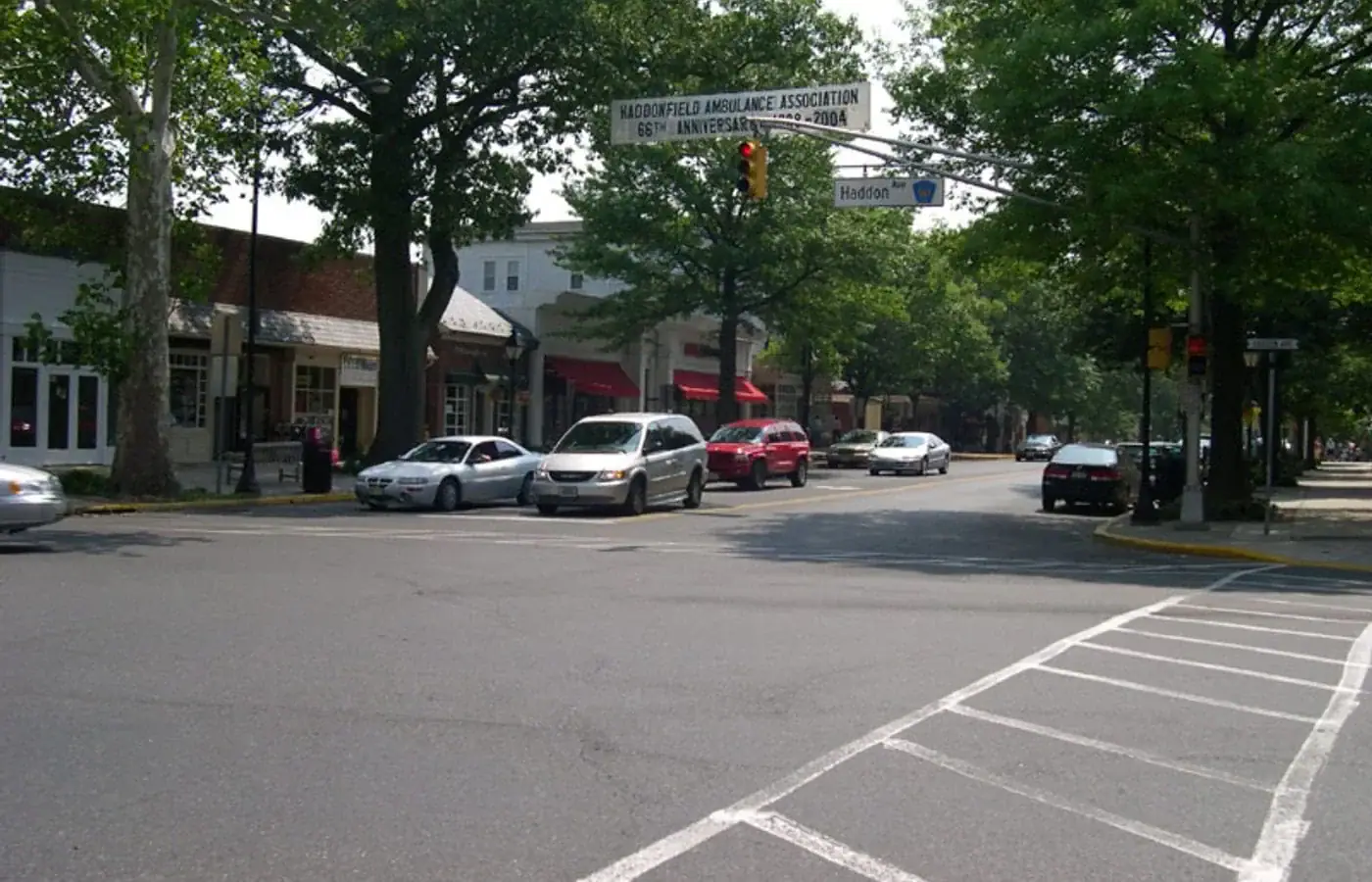 Street-level view of a town intersection in a Gloucester County painting, featuring cars and a banner reading Haddonfield Ambulance Association 66th Anniversary 1938-2004.