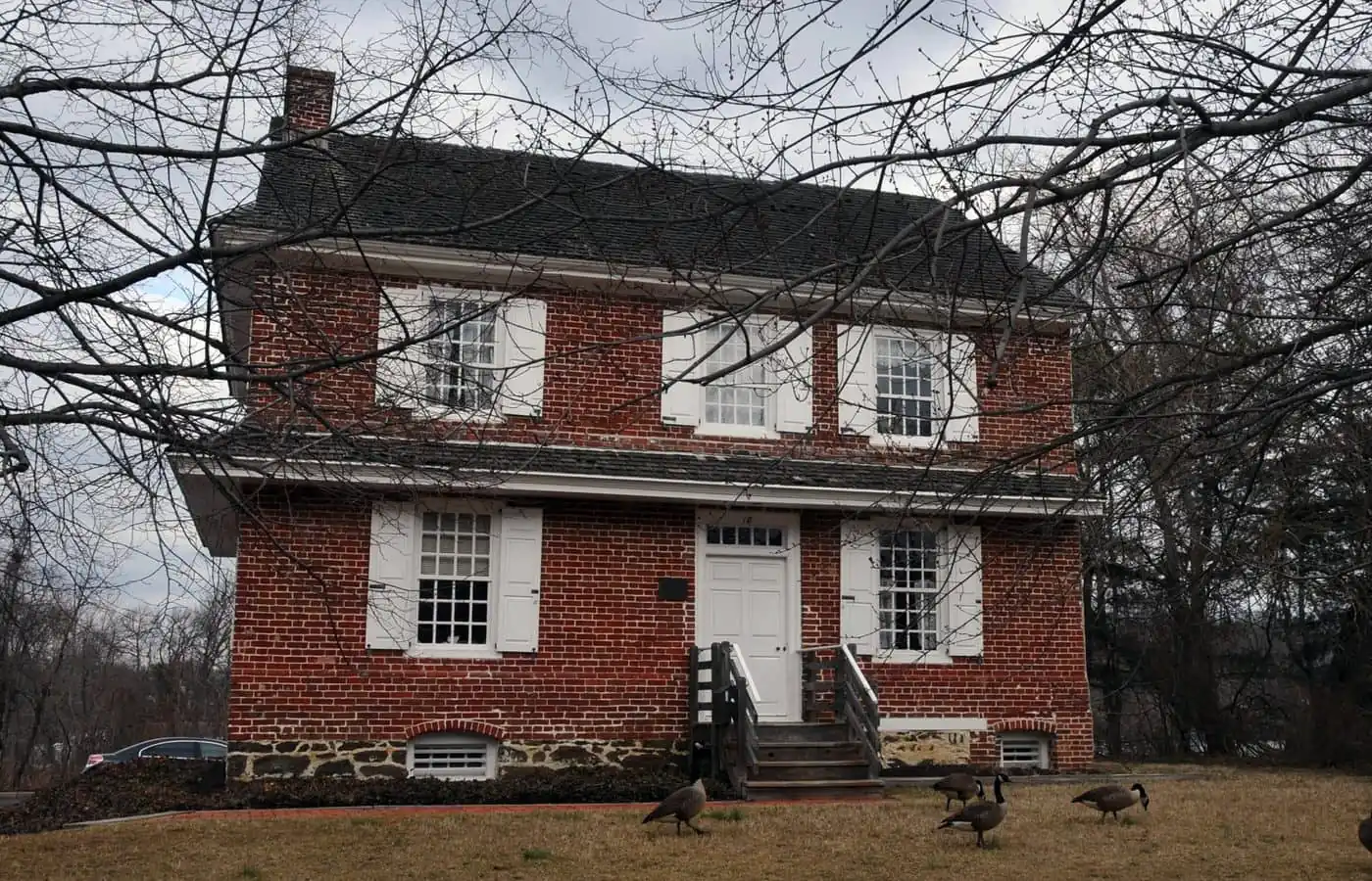 A two-story red brick house with white shutters and a central white door, featuring the work of Camden County painters, with four geese walking on the lawn in front.