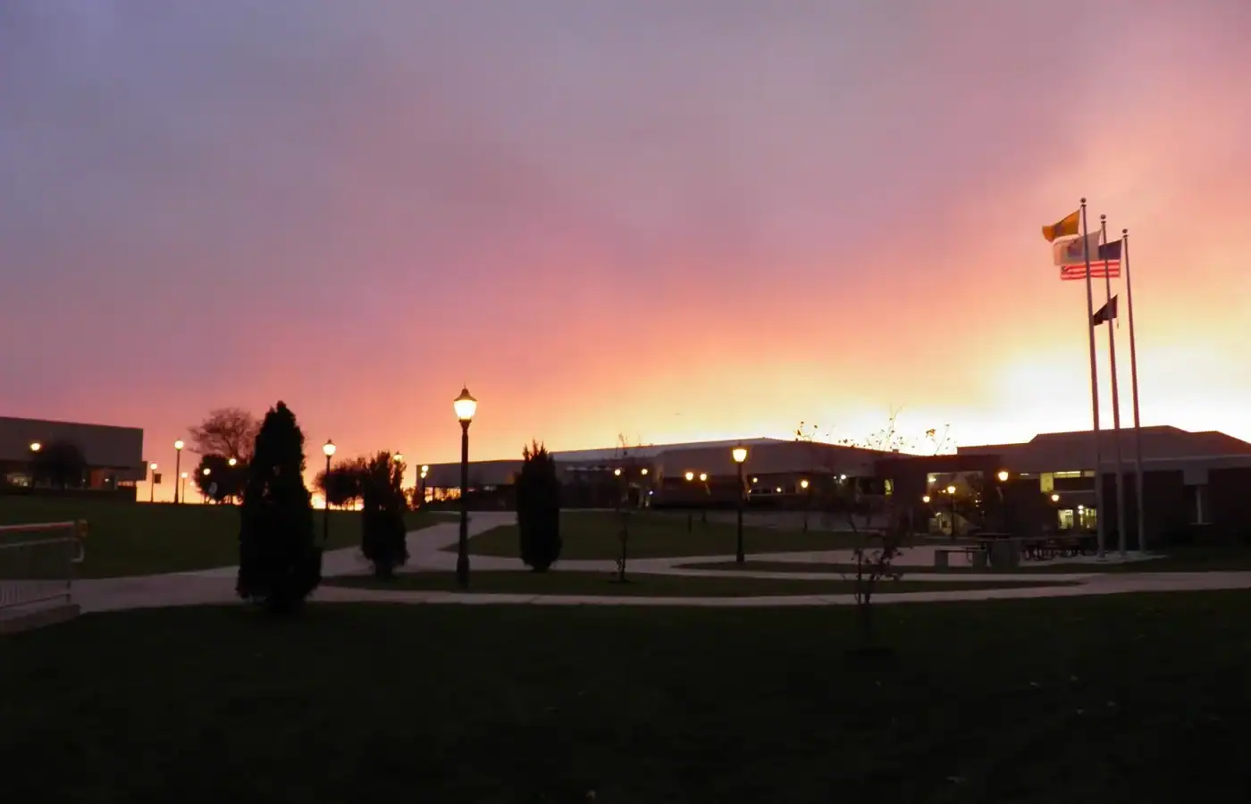 A school campus at sunrise in a Gloucester County painting, with buildings silhouetted against a purple and orange sky, featuring a row of lit lamp posts and three flags on poles.